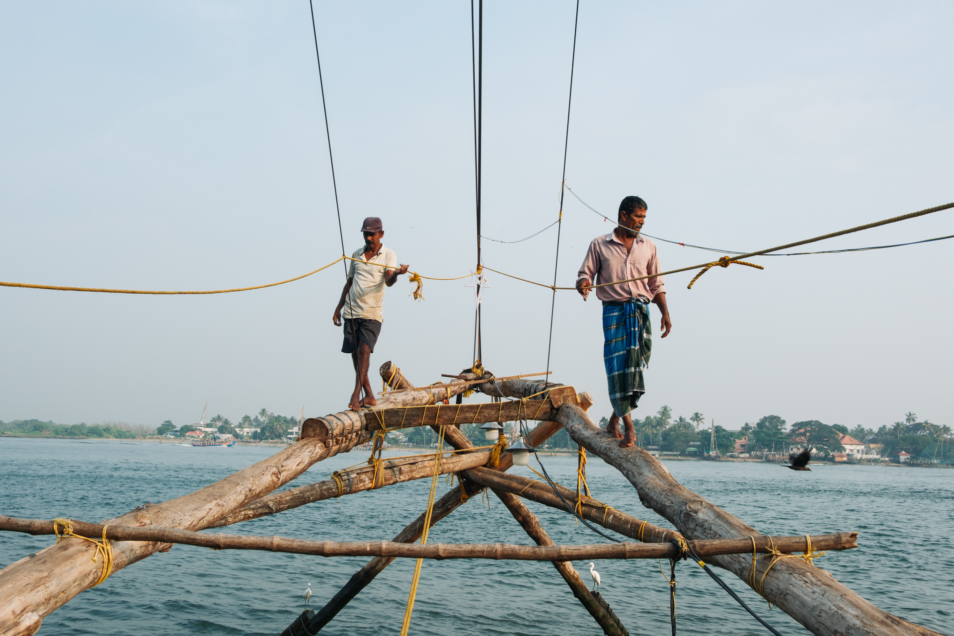 Two fishermen balancing on wooden beams as they operate a traditional Chinese fishing net over the water in Fort Kochi, Kerala.