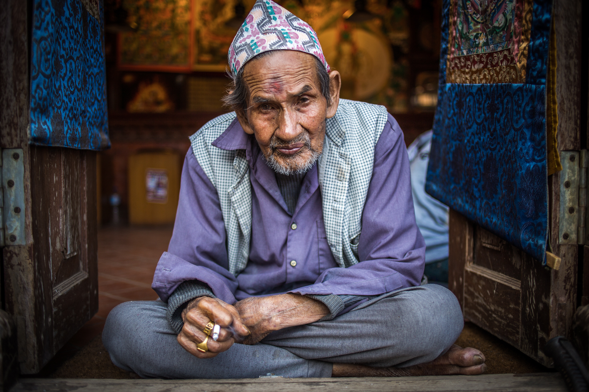 An old Thangka painter from Bhaktapur, Nepal, is smocking a cigarette on the entry of his shop. The hat on his head is called