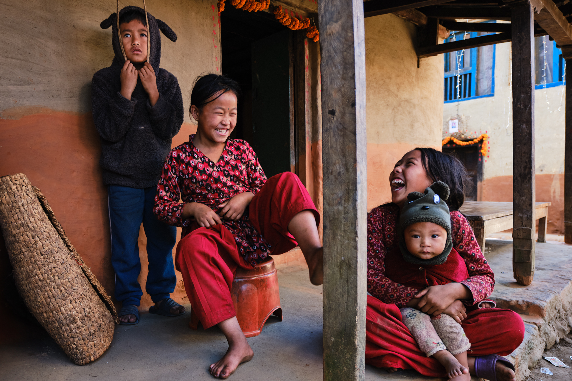 Nepali children laughing and playing on a house porch during Tihar, wearing red clothes and surrounded by warmth and joy.
