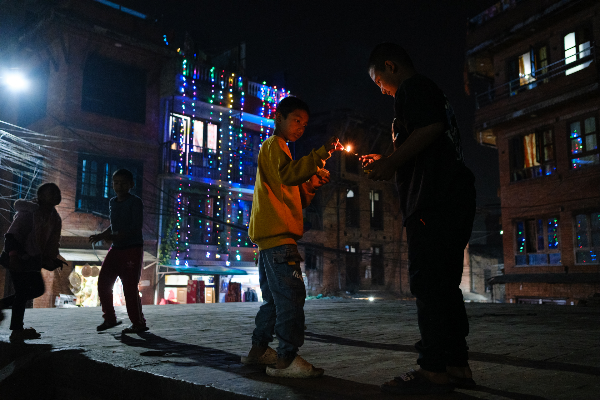 Two children lighting a sparkler under festive lights during Tihar evening in a Nepali courtyard
