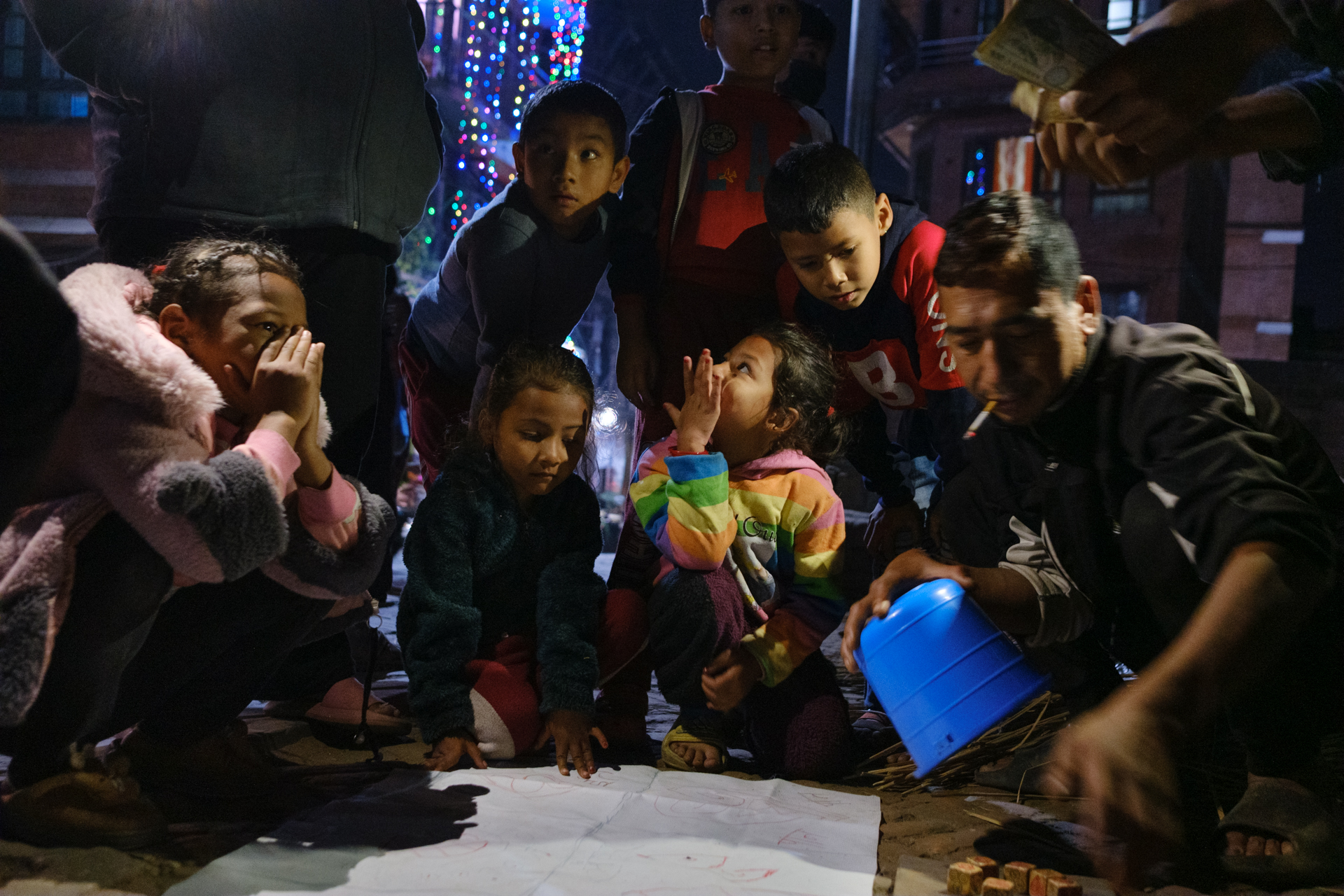 Group of children and adults gathered at night playing a traditional street game during Tihar, lit by streetlights and fairy lights.