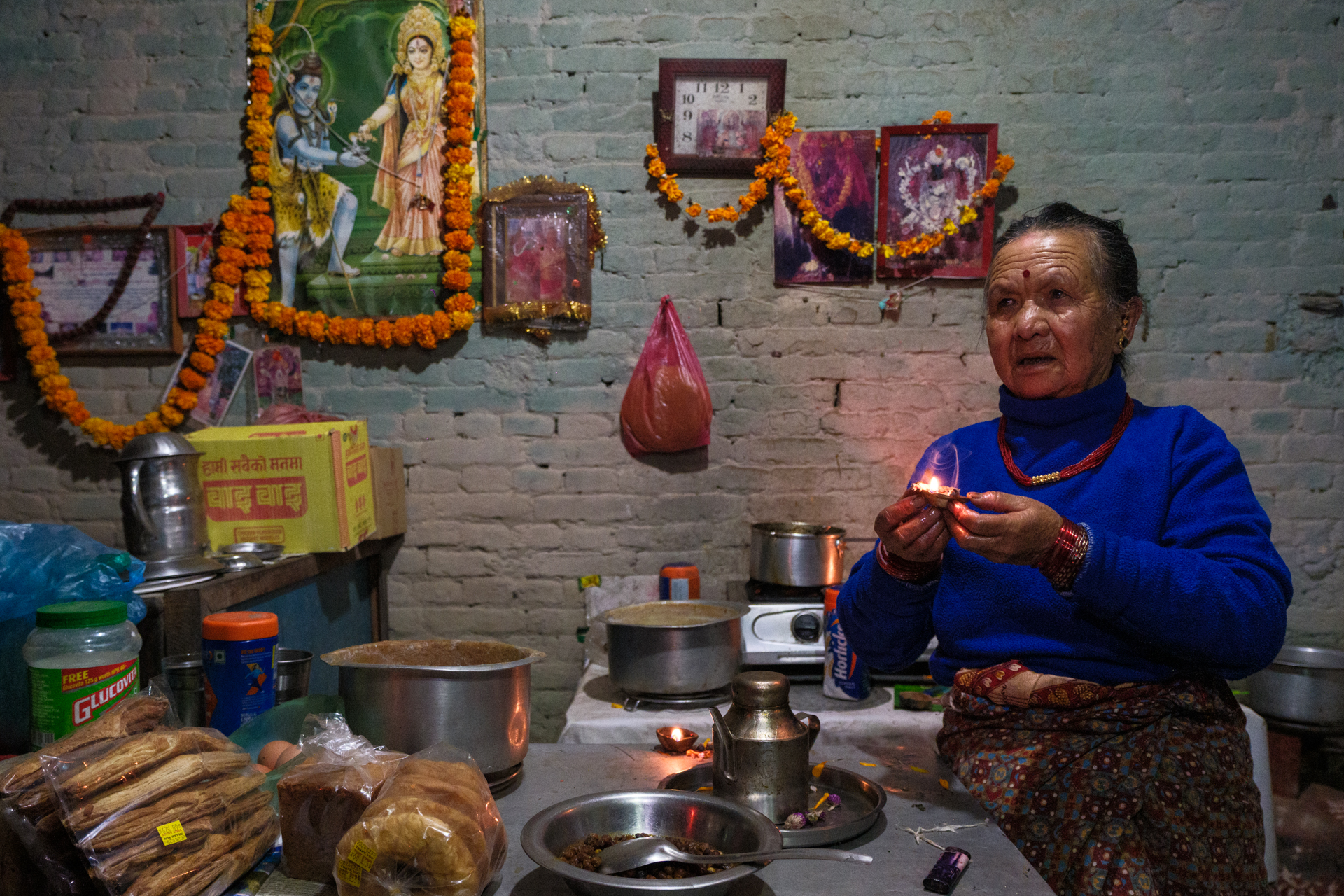 Elderly Nepali woman holding a burning oil lamp in her kitchen during Tihar, with devotional posters and food offerings around her