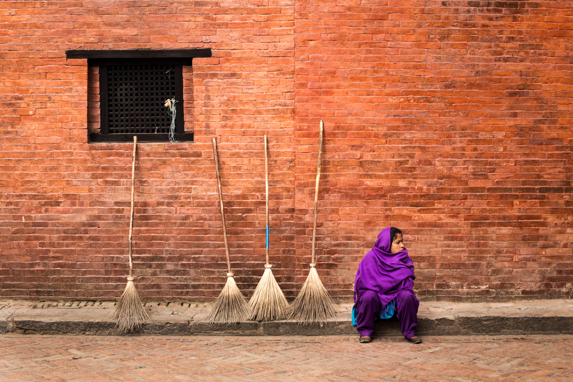 A woman in a purple shawl, is sat in the old streets of Kathmandu, near 4 broomsticks that she is selling. Nepal photography by Lorenz Berna