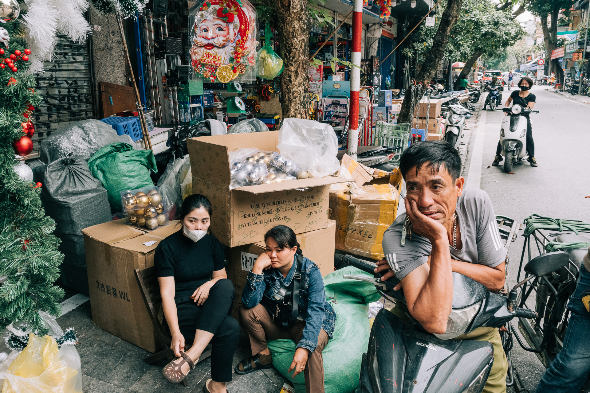 street photo of People resting and chatting among boxes and goods on a busy Hanoi street market.