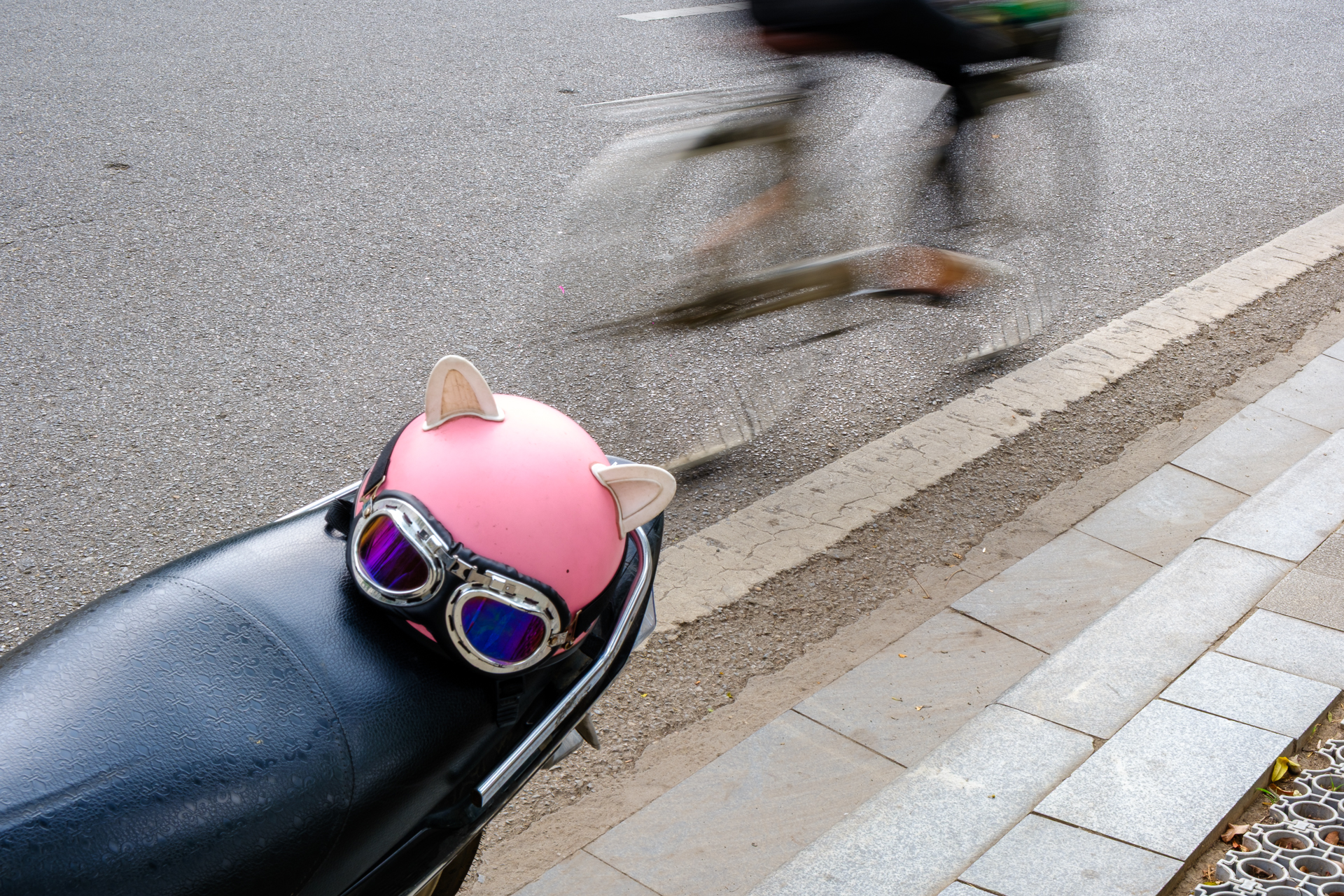 A scooter with a pink cat-shaped helmet parked as a cyclist rides past in motion blur.