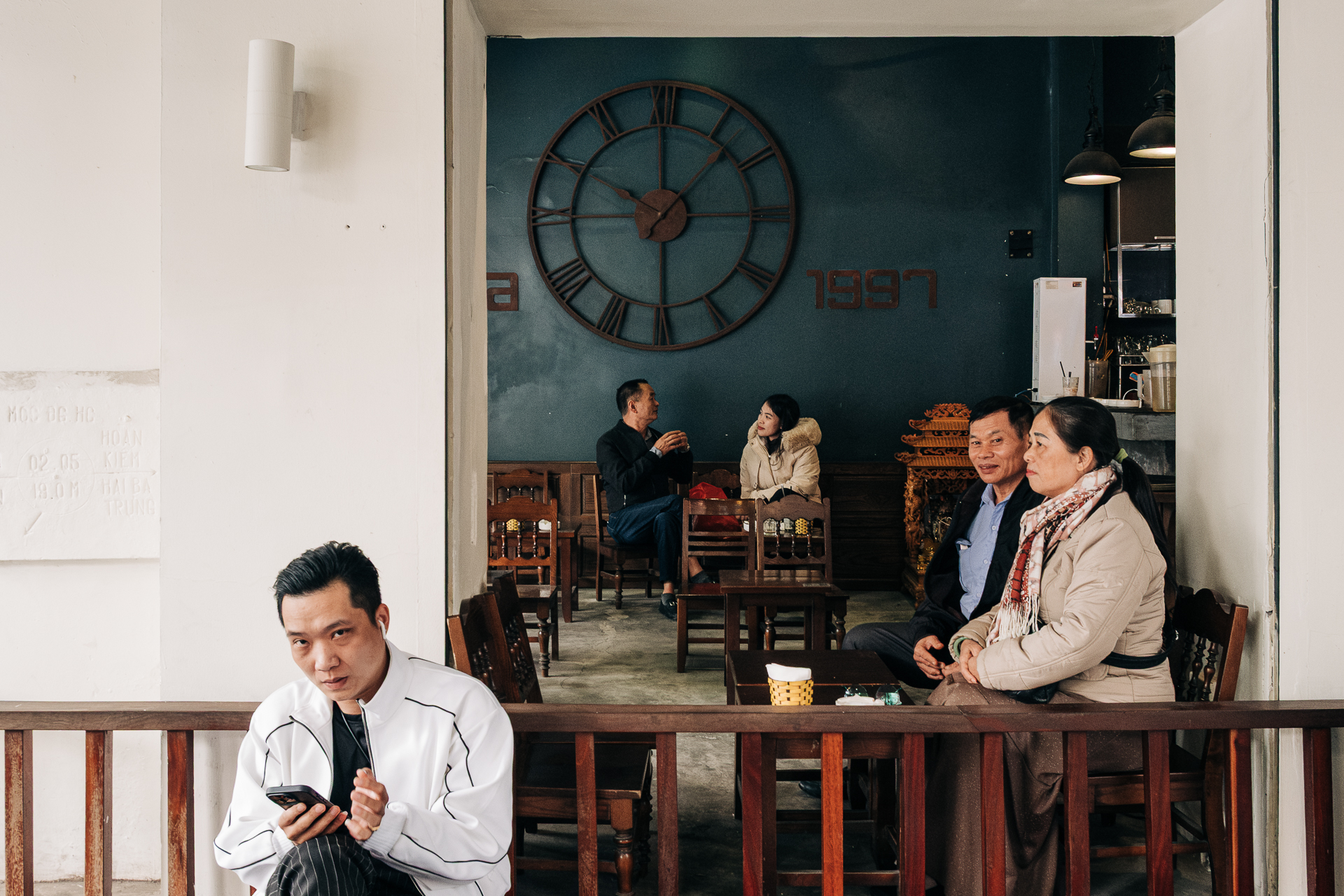Young man sitting outside a Hanoi café, with people gathered and a large clock visible inside.