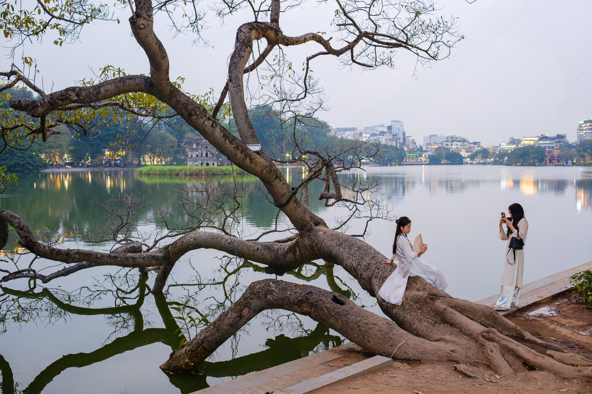 Two women posing and taking photos on a large tree beside Hoan Kiem Lake in central Hanoi.